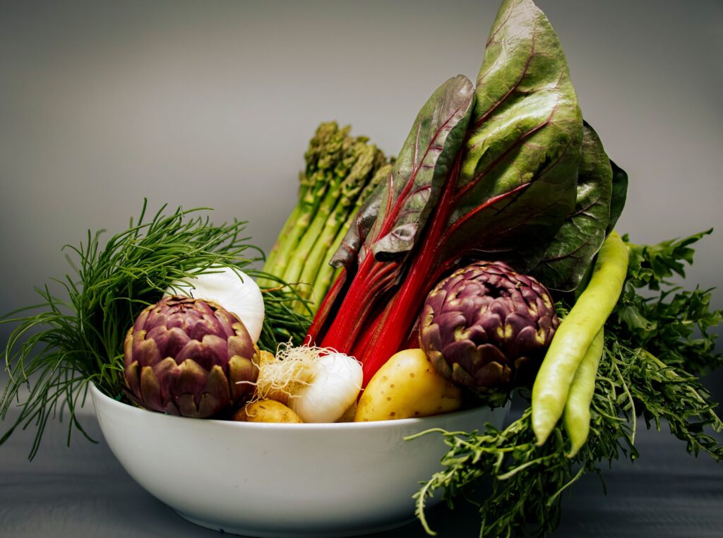 A vibrant assortment of fresh vegetables including artichokes, chard, and asparagus in a white bowl.