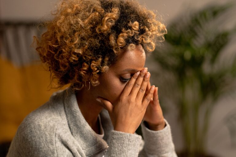 A woman with curly hair in a gray sweater, holding her face, depicting stress indoors.