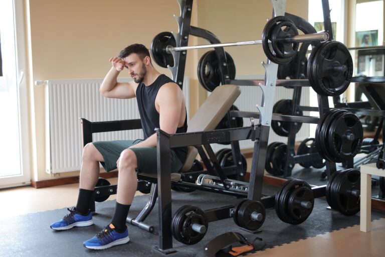 Man taking a break in a gym setting, sitting beside loaded weight bench, looking thoughtful.