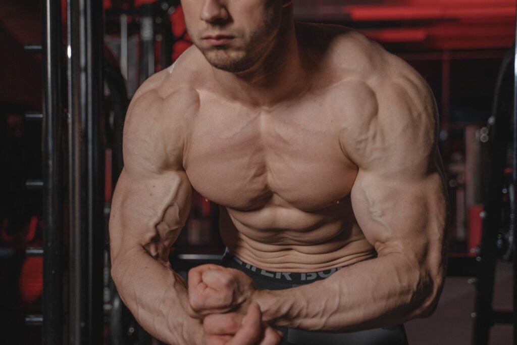 Shirtless man displaying muscular physique with focus on biceps and abs in a gym.