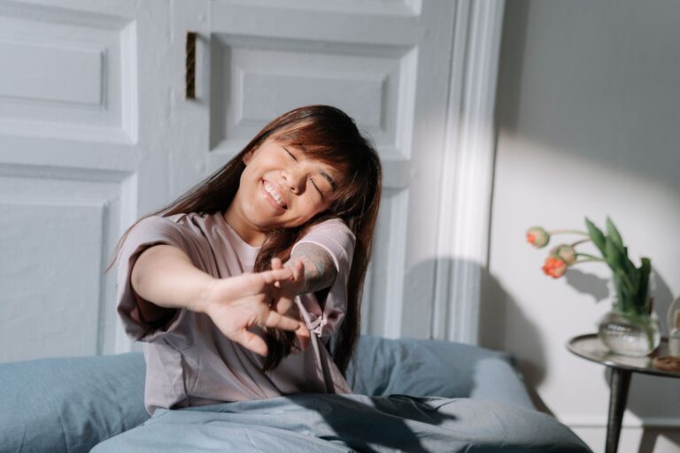 Smiling Asian woman stretching in bed, enjoying soft morning light indoors.