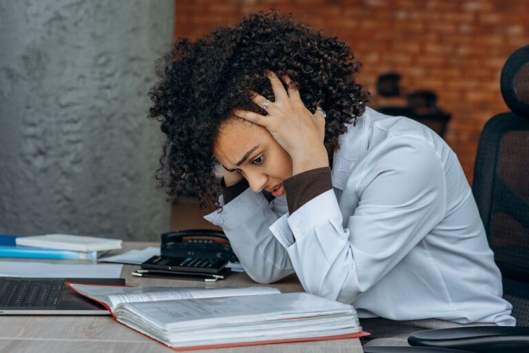 Woman sitting at desk appearing stressed while reviewing documents in an office setting.