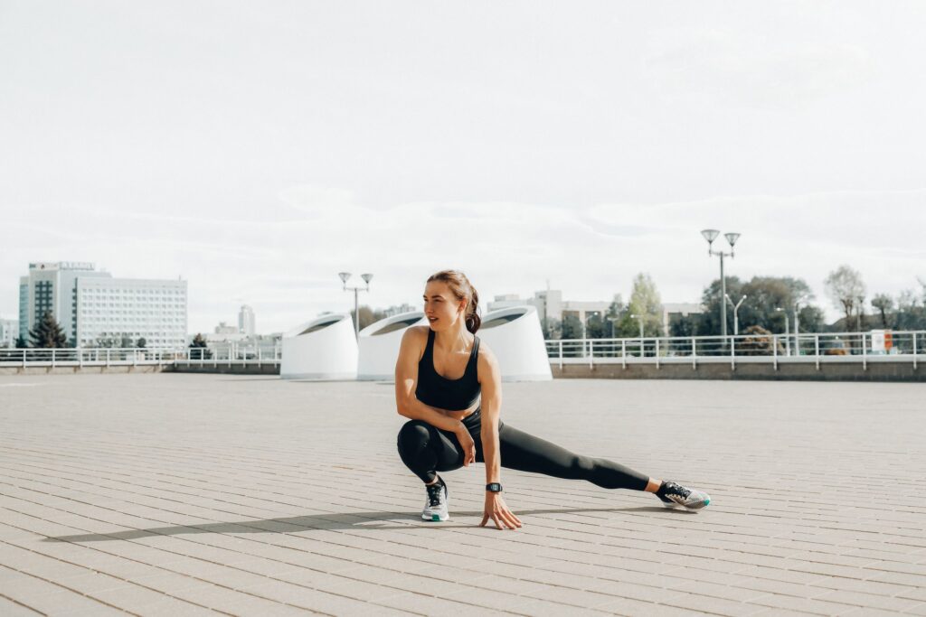 Young woman in black sportswear stretching outdoors in a sunny urban environment.