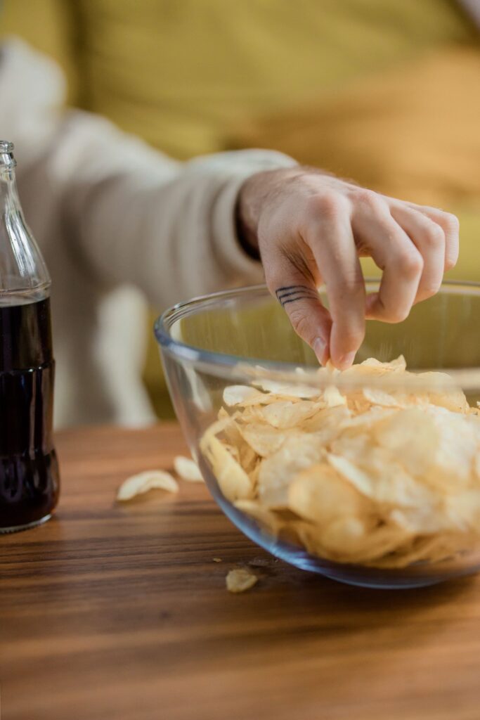 A close-up of a hand reaching for potato chips in a bowl, beside a soda bottle.