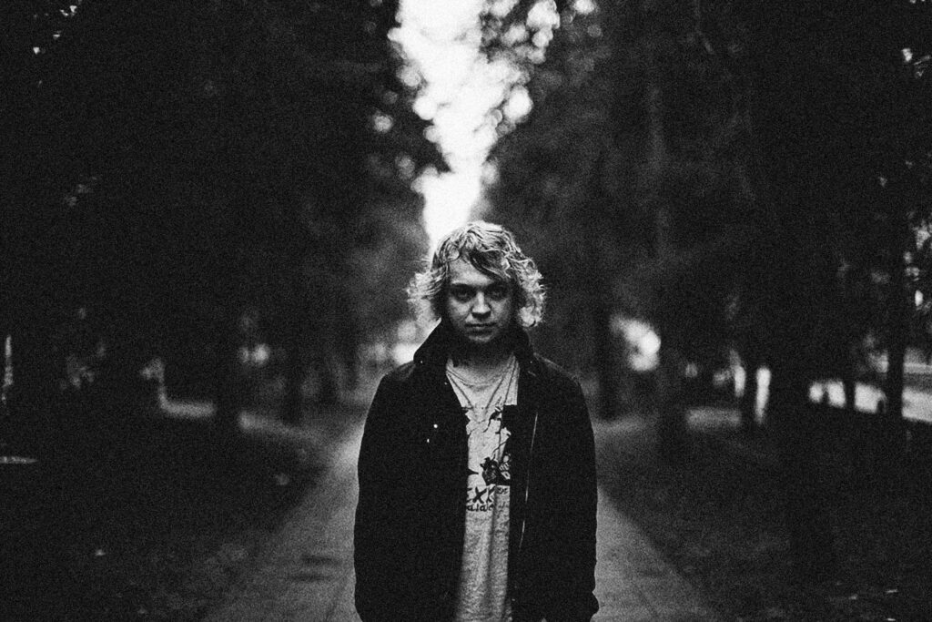 A moody black and white portrait of a person standing on a tree-lined path in an urban park.