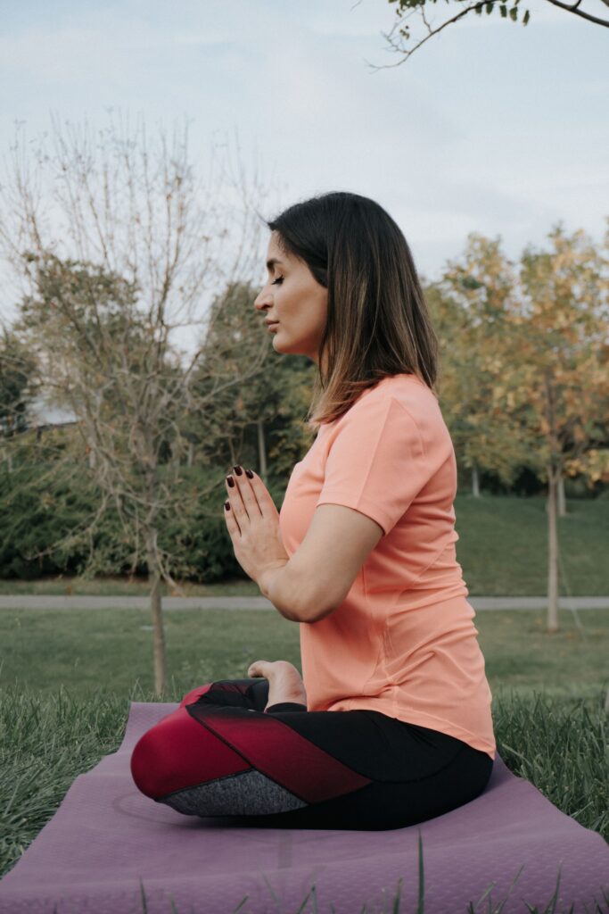 A woman meditating on a yoga mat outside in a park during the day.