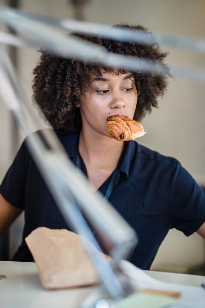 An adult woman with curly black hair enjoys a pastry at her office desk, multitasking during work hours.