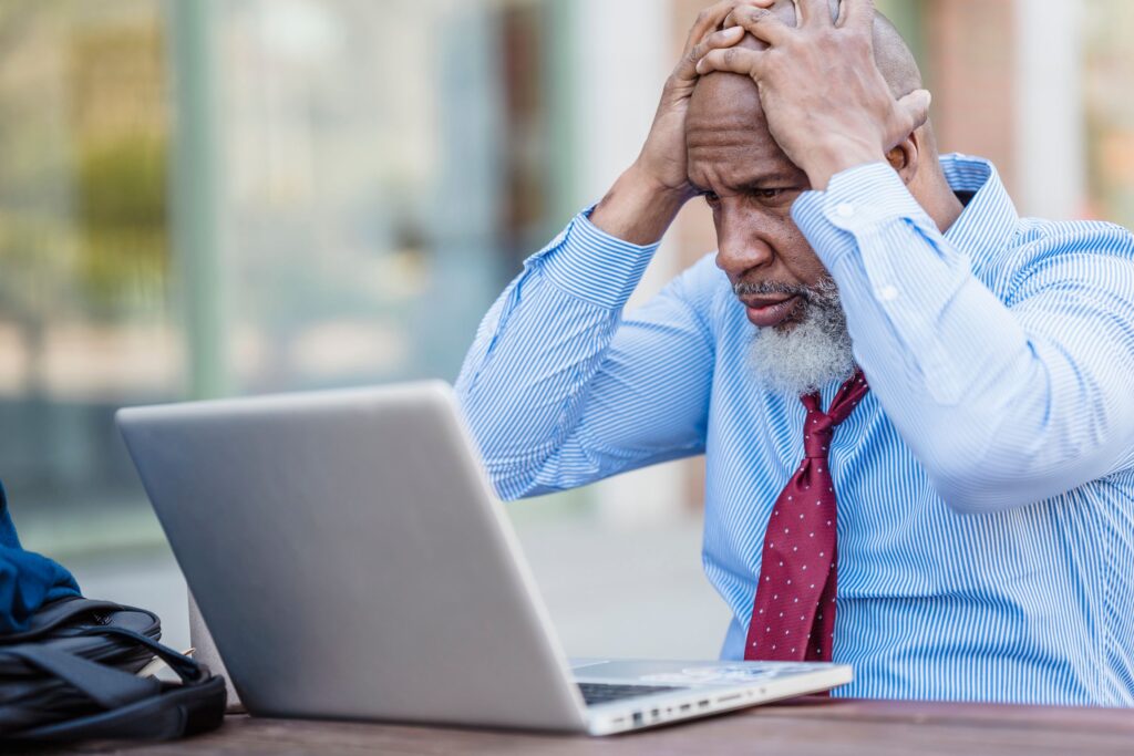 Anxious senior African American male entrepreneur in formal classy shirt and tie sitting at table with laptop and grabbing head