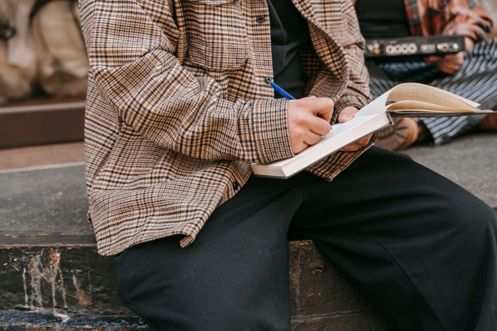 Close-up of a person writing in a notebook with a pen, sitting outdoors on a sunny day.