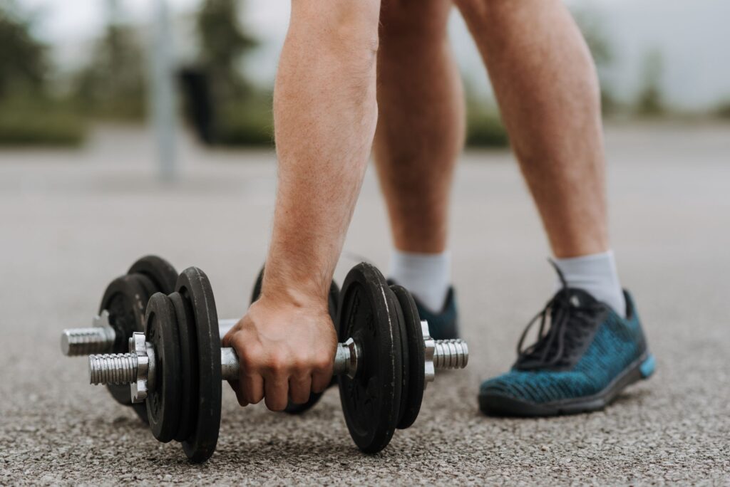 Crop anonymous strong male lifting heavy metal dumbbell placed on asphalt road in soft focus