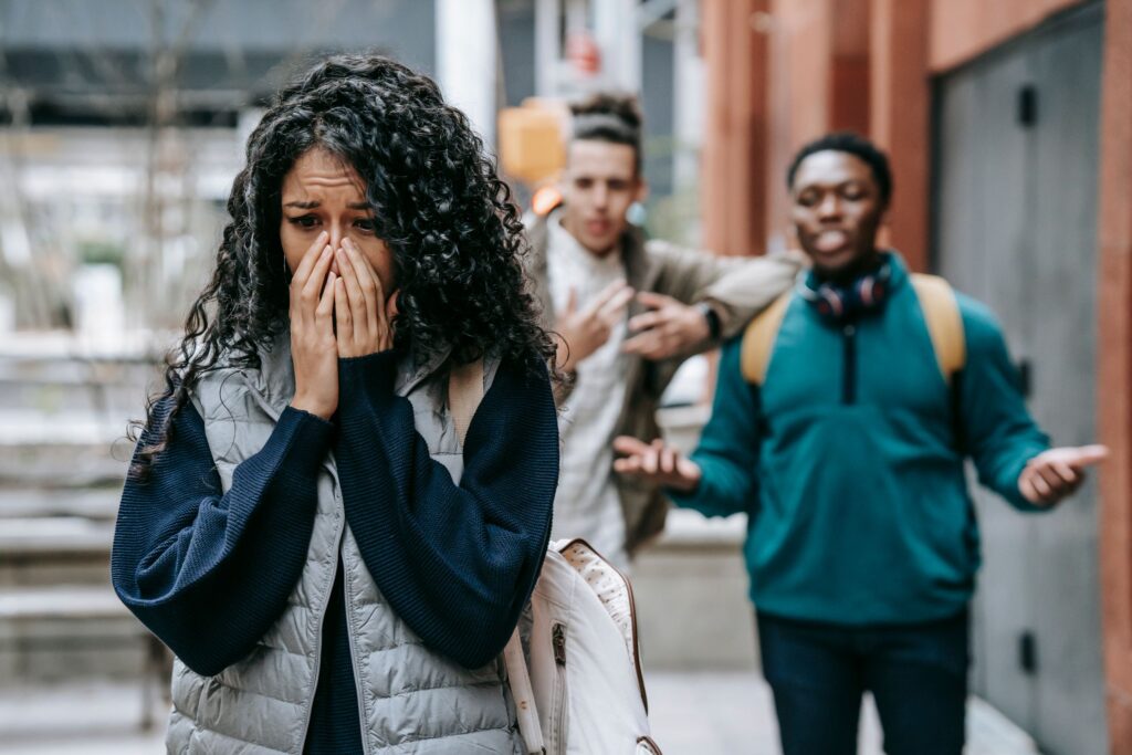 Diverse young male friends scoffing at ethnic female groupmate crying and covering mouth with hands on street