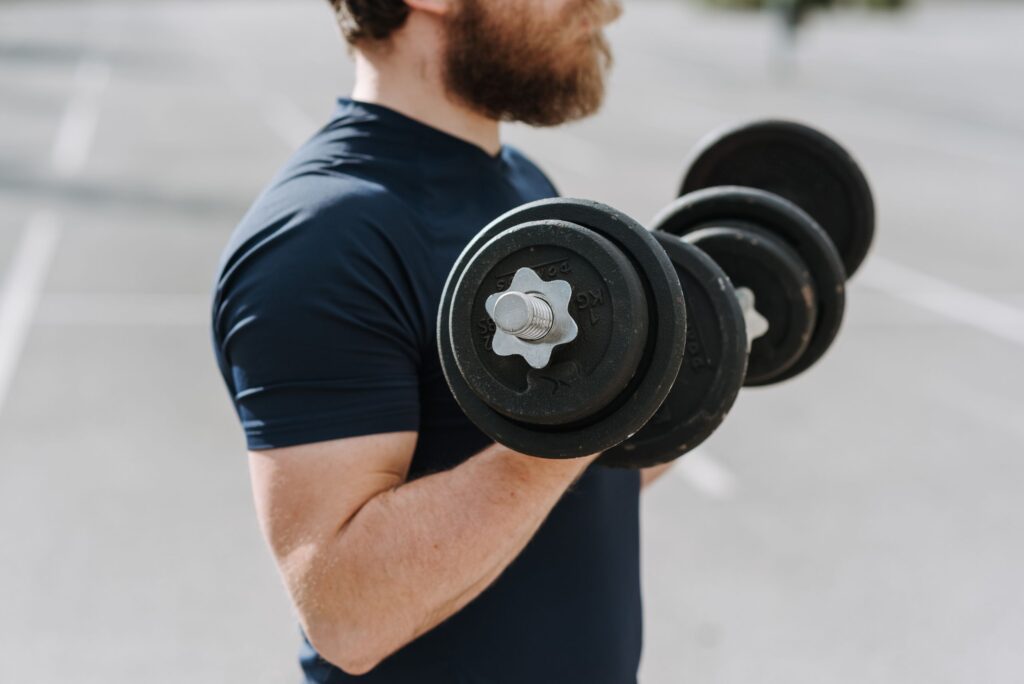 Side view of crop male in t shirt doing bicep curls with dumbbells training on sports ground