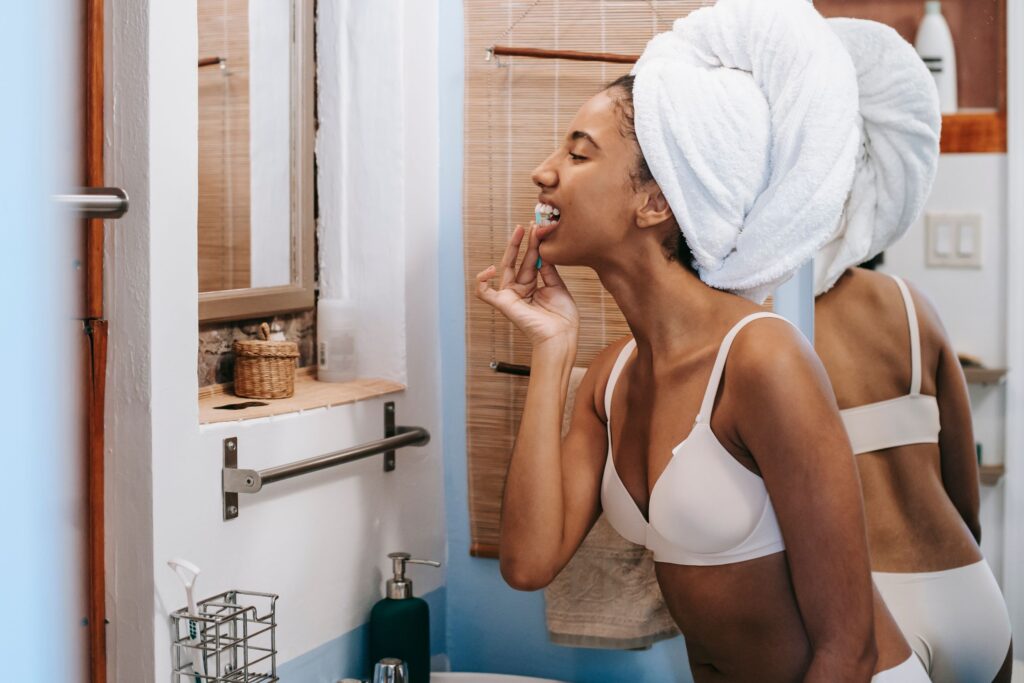 Side view of young African American female in underwear with white towel on head standing in front of mirror in bathroom and cleaning teeth with dental flosser