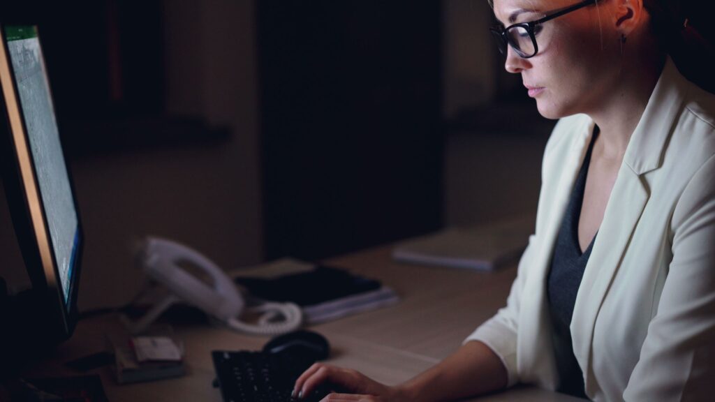 Woman in glasses working late at a desk with a computer monitor.