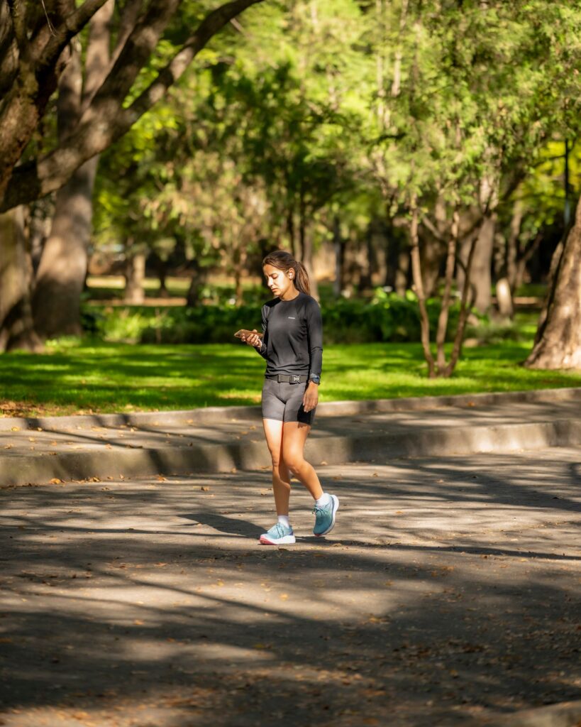 Woman jogging in a sunlit park, checking smartphone while exercising outdoors.